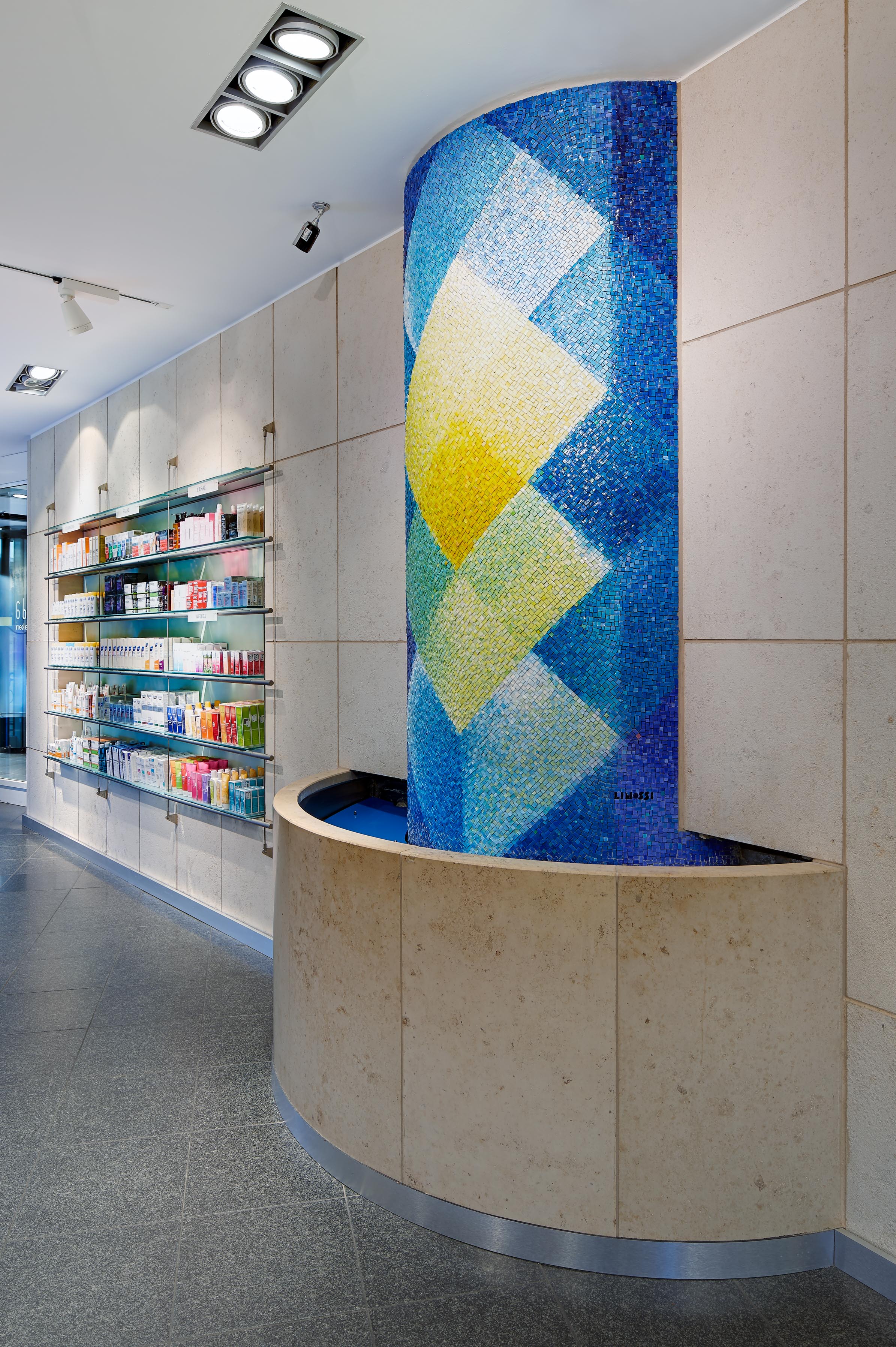 Detailaufnahme des halbkreisförmigen Wandbrunnens mit dem farbenprächtigen, blau-gelben Mosaik des Künstlers Lino Linossi