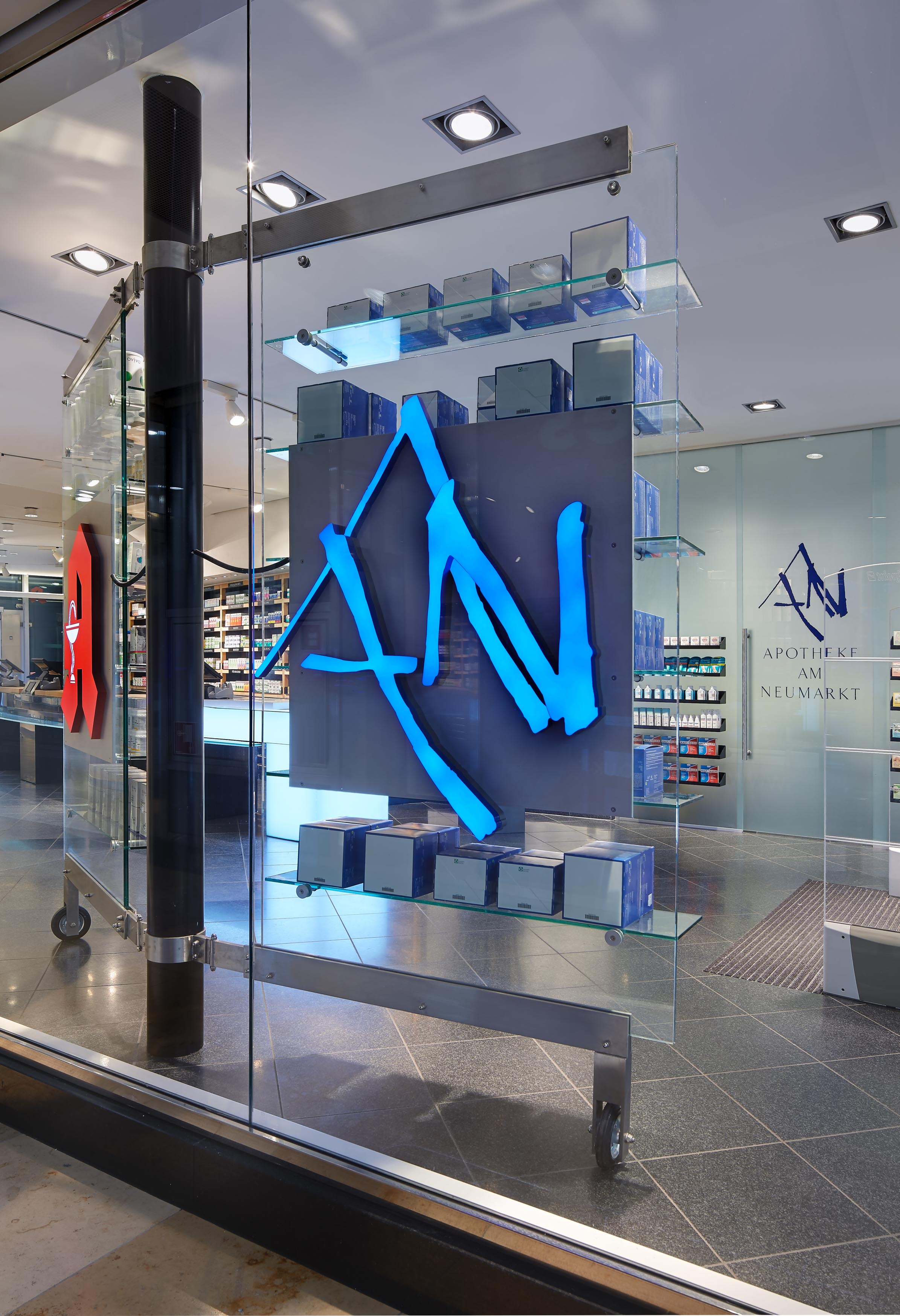 Nahaufnahme des blau leuchtenden, stilisierten AN-Logos der Apotheke auf einem freistehenden Glasregalsystem im Schaufenster