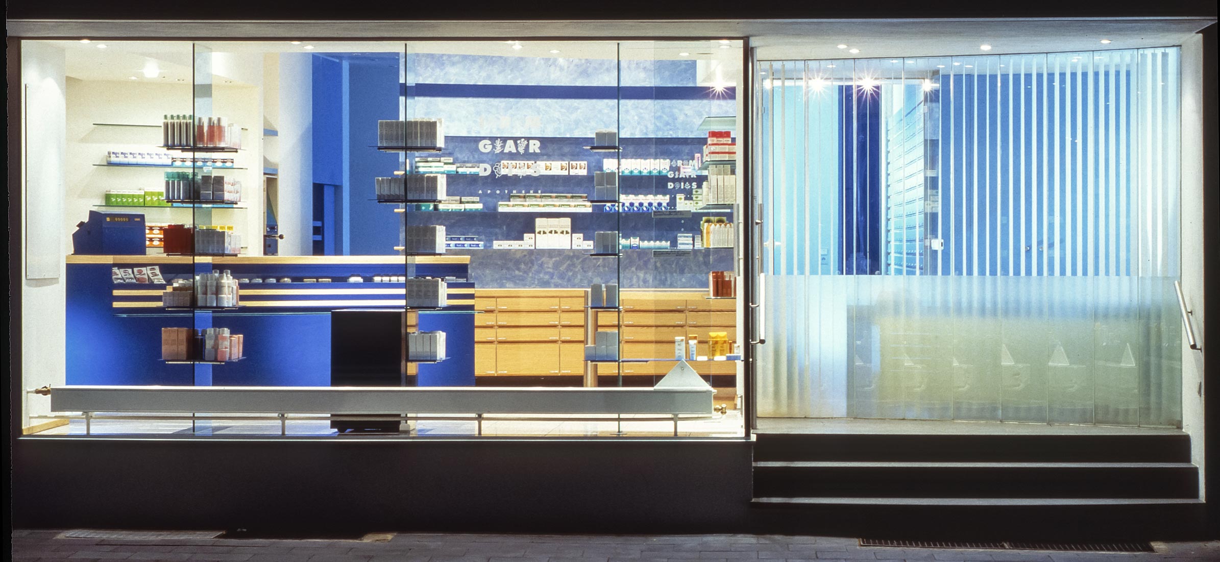 Großflächige Schaufensterfassade der Irmgardis-Apotheke bei Nacht mit Blick auf das blaue Interieur